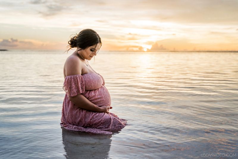 pregnant woman in flowy pink dress sitting in water with sunset on background